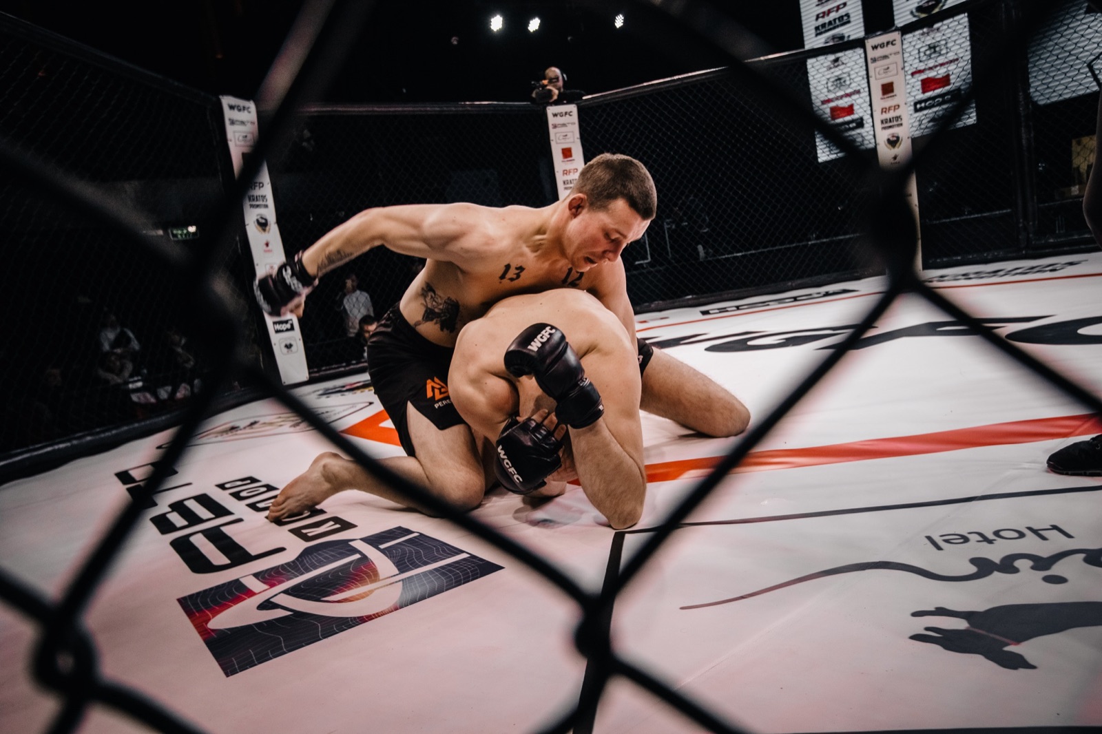 A fighter controls an opponent on the ground, seen through the cage mesh.