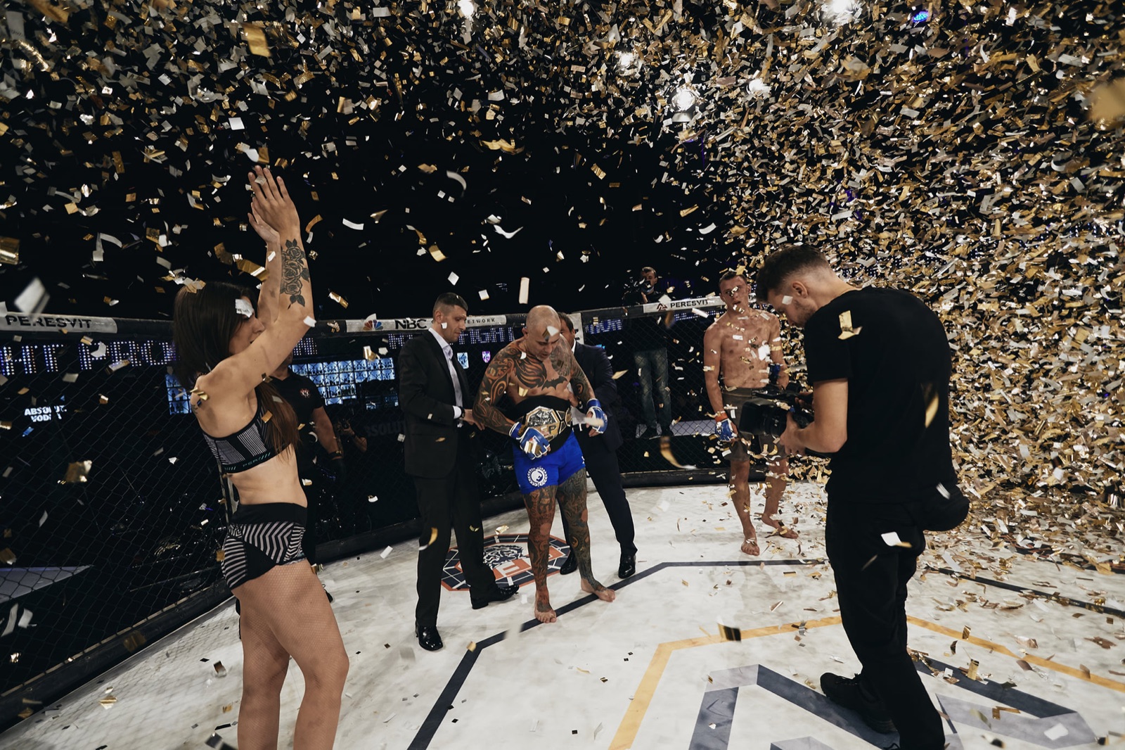 A champion wearing the belt stands inside the cage under falling gold confetti.