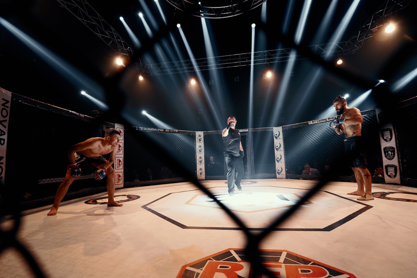 Two fighters begin an exchange under theatrical beams of light, seen through the cage.